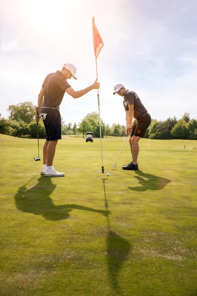 Zwei Golfer in dunkler Kleidung beim Putten auf sonnigem Gr&uuml;n, Flagge im Loch, B&auml;ume und Golfcart im Hintergrund