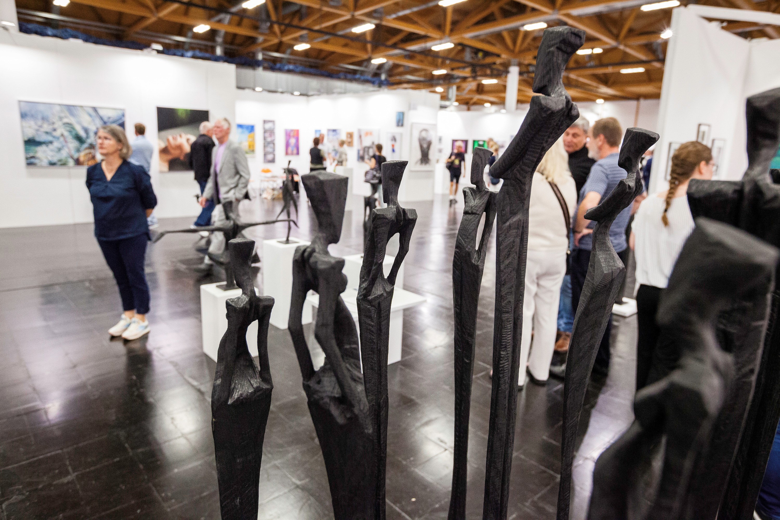 Schwarze abstrakte Skulpturen im Vordergrund, Besucher betrachten Gemälde und Skulpturen auf Kunstmesse in heller Halle
