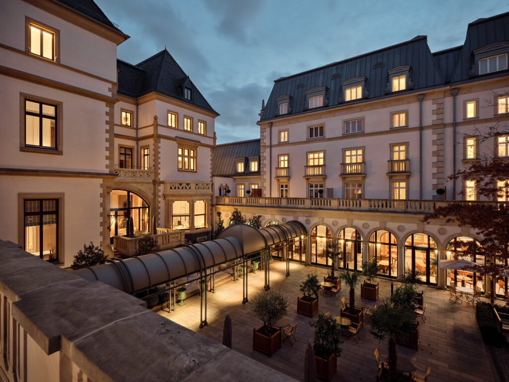 Historische Fassade eines Hotels mit beleuchtetem Innenhof, Glas&uuml;berdachung und Sitzbereichen am Abend