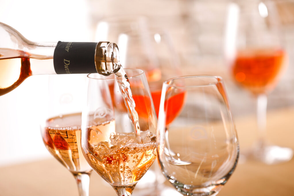 Roséwein wird in ein Probierglas eingeschenkt, mehrere gefüllte Weingläser im Hintergrund auf Holztisch