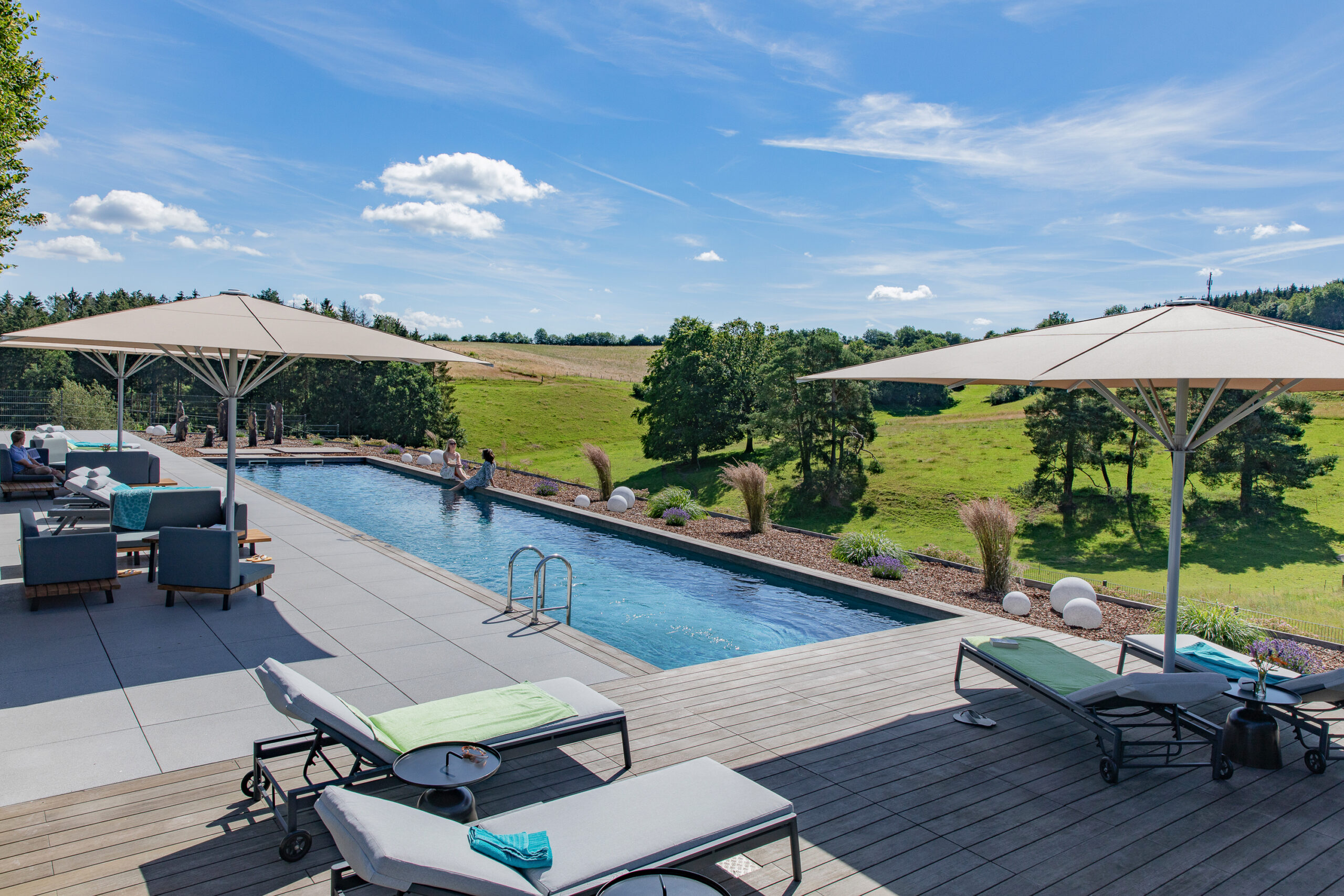 Modernes Ferienhaus mit langem Außenpool, Liegen und Sonnenschirmen, Blick auf grüne Wiesen und Bäume in der Eifel