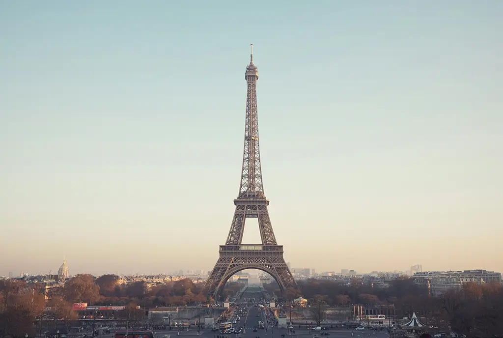Der Eiffelturm in Paris, zentral vor einem leicht bewölkten Himmel, umgeben von Gebäuden und Bäumen im Vordergrund.