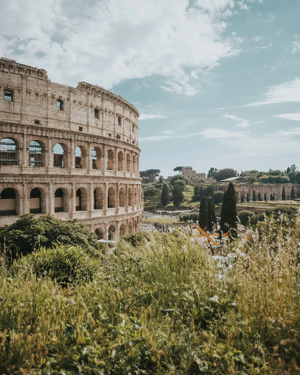 Das Kolosseum in Rom, Italien, umgeben von grünen Pflanzen und einer weiten Landschaft unter einem teils bewölkten Himmel.